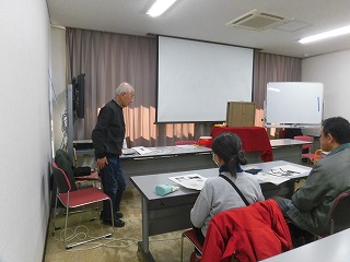 武さんがプロジェクターのスクリーン前で立ち、テーブルを囲んで座る少人数の参加者に説明をしている様子の写真