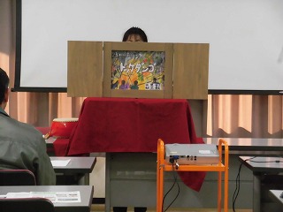 白いスクリーンと赤布がかけられた演台の前で、一人の女性が「トッケダンコ」と書かれた紙芝居を披露しており、観客の一人がその様子を椅子に座って見守っている様子の写真