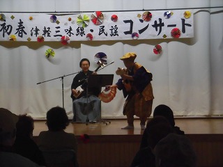 白い壁に「初春津軽三味線・民謡コンサート」と書かれた横断幕が掲げられた舞台の上で、津軽三味線を演奏する男性と、拍子木を叩いて踊りを披露する人物を観客が鑑賞している写真