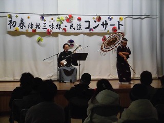 白い幕が背景のステージで、和装の二人が津軽三味線の演奏と傘踊りを披露しており、「初春津軽三味線・民謡コンサート」と書かれた横断幕が掲げられている写真