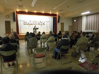 大勢の観客が椅子に座ってステージに注目しており、和装の二人が「初春津軽三味線・民謡コンサート」と書かれた幕の前で演奏と踊りを披露している様子の写真