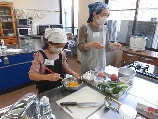 家庭科室で、エプロンとバンダナを身につけた親子が、目の前に並べられた食材を使い、ボウルの中で卵を混ぜながら楽しそうに料理教室に参加している写真