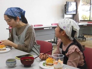家庭科室のテーブルで、親子が料理教室で作ったちらし寿司やデザートを前に、お互いの料理を見ながら楽しそうに食事をしている写真
