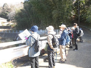 様々な帽子とリュックを背負った男女のグループが、冬枯れの景色の中、白いガードレール越しに川を覗き込みながら、手に持った資料を熱心に見ている様子の写真