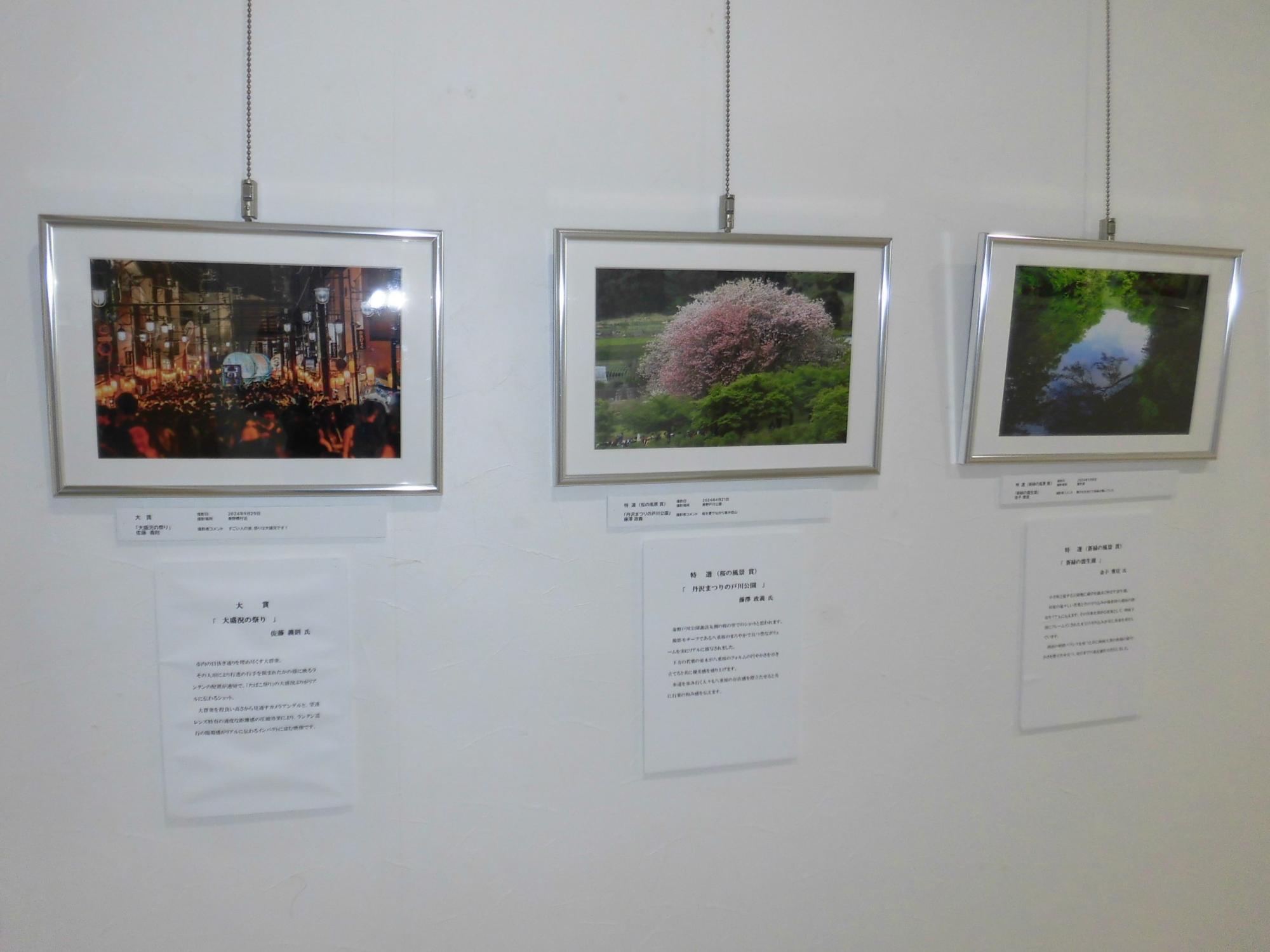 白い壁に3枚の額縁入り写真が展示され、それぞれにキャプションが添えられており、夜祭の風景や新緑、満開の花といった自然や文化の作品が並ぶ写真