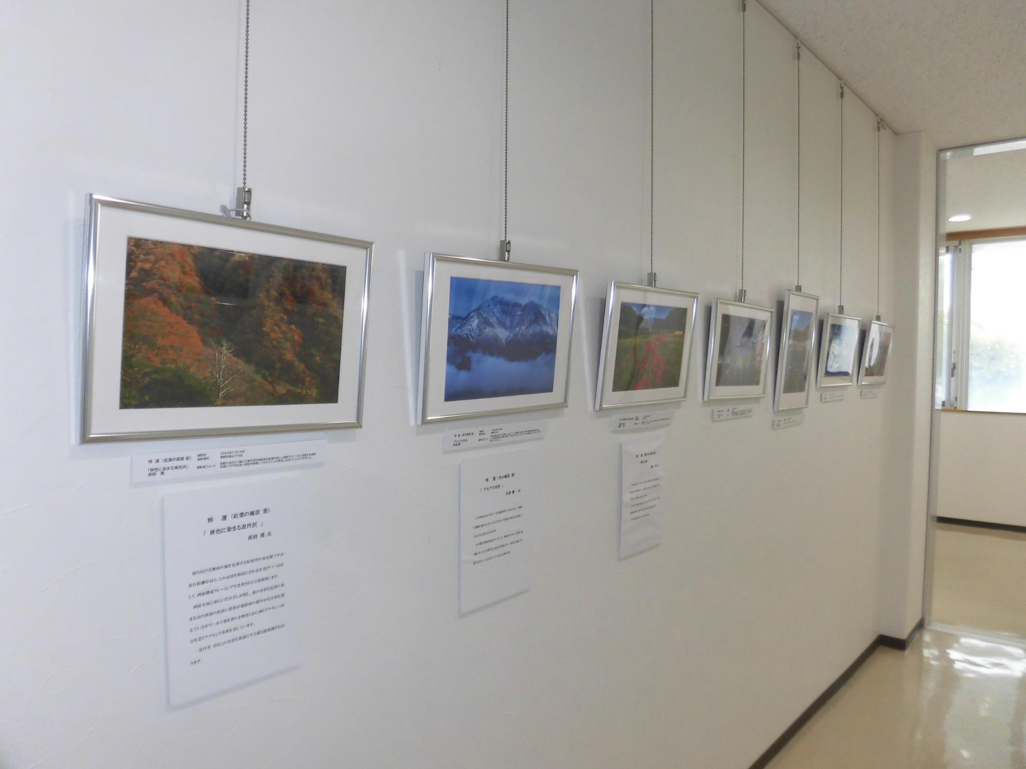 白い壁に複数の額縁に入った写真が展示されており、それぞれにタイトルと説明文が添えられている、紅葉や雪山、川沿いの風景をテーマにした写真展の様子の写真