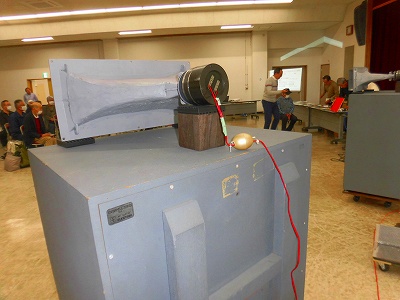 講義室のような屋内で、手作りの複雑なスピーカーシステムが展示されており、多くの参加者たちが会場の席に座っている様子が見えるレコードカフェ「メモリーズ」の様子の写真