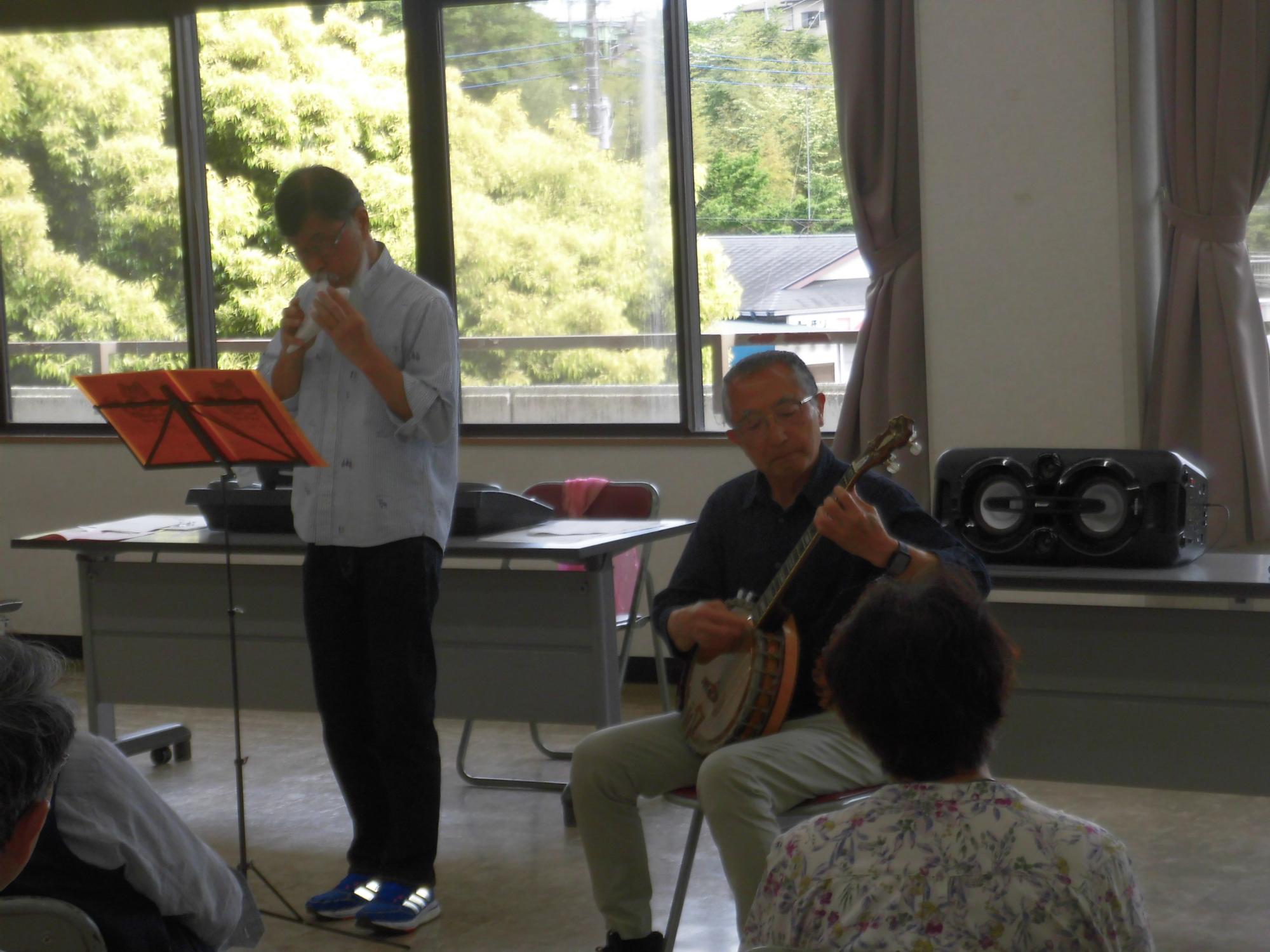屋内広間にて、一人の男性がバンジョーを演奏し、もう一人の男性が譜面台の楽譜を見ながらハーモニカを吹いている、音楽発表会の様子を写した写真