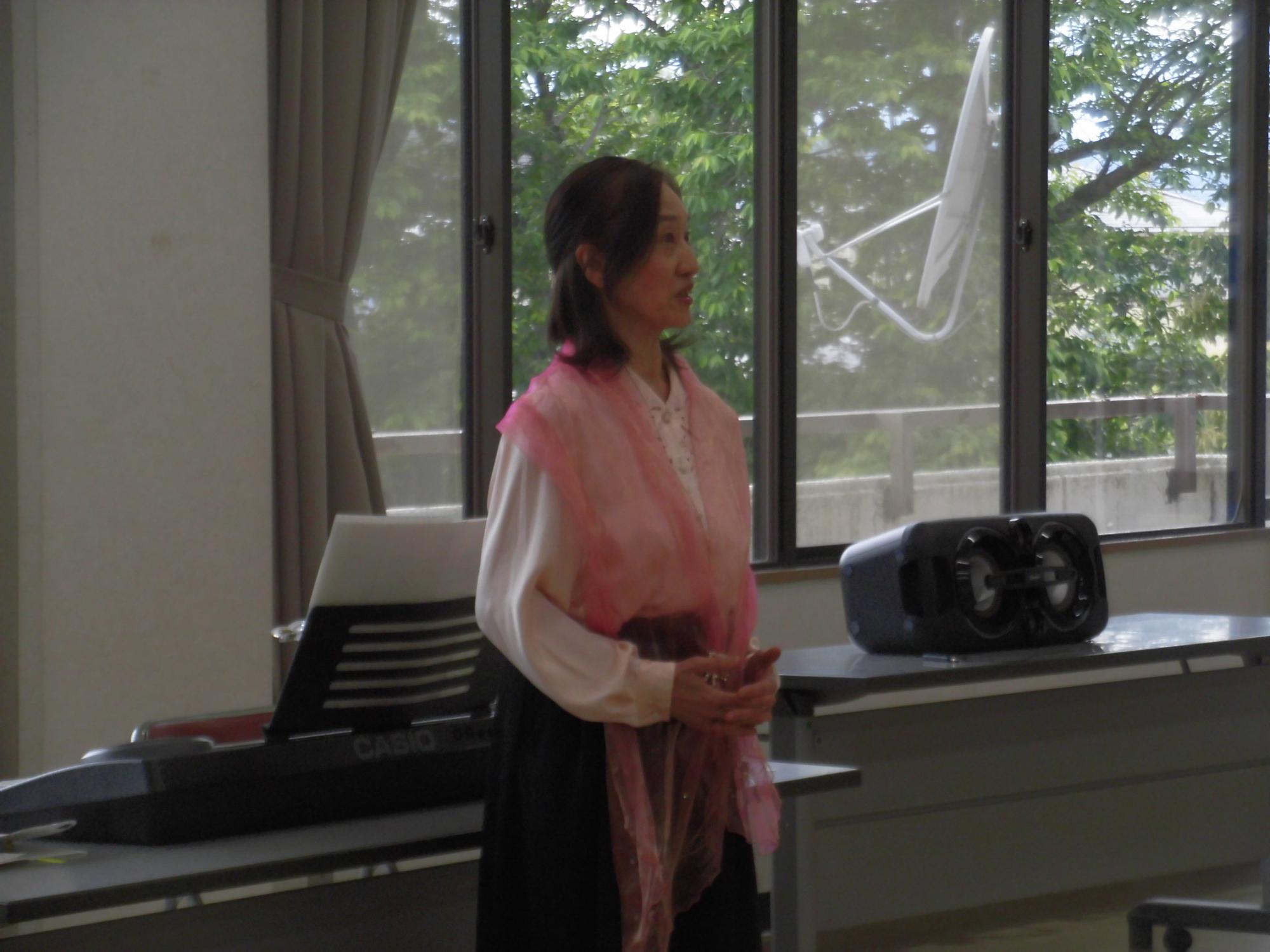 マイクに向かって話す女性がピンク色のストールを首に巻いており、背景には電子キーボードやスピーカー、大きな窓が見える講演会の様子を捉えた写真