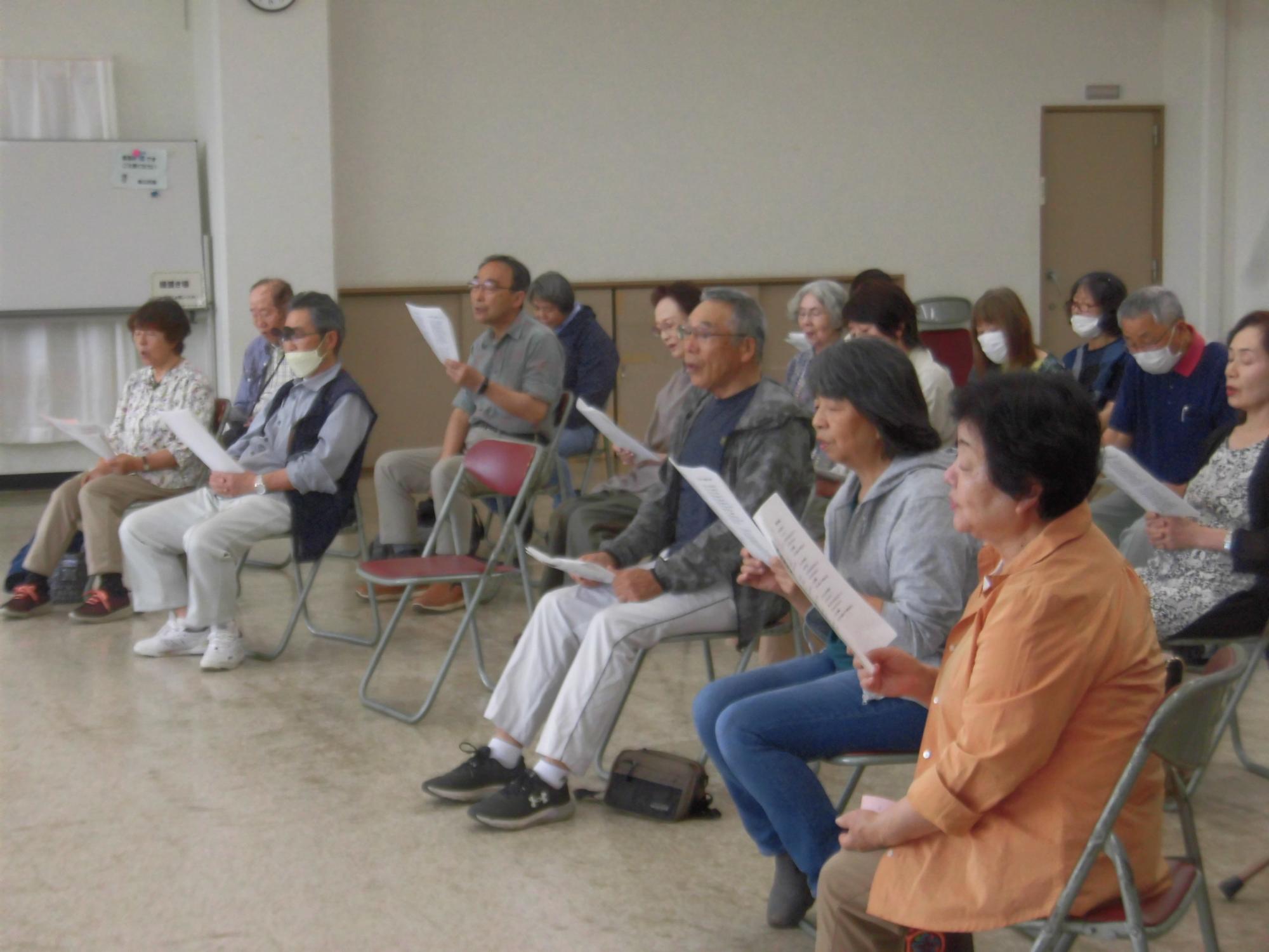 大勢のシニア世代が、椅子に座ってそれぞれ譜面や歌詞が書かれた紙を持ちながら、熱心に合唱練習に取り組んでいる様子の写真