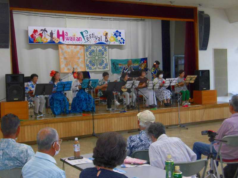 屋内のステージで、ハワイアンフェスティバルの横断幕を背景に、民族衣装を着た複数の男女がウクレレを演奏し、観客がそれを見ている様子を捉えた写真