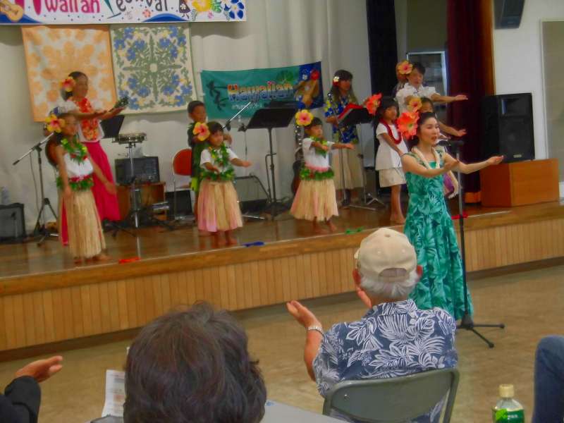 Hawaiian Festivalの看板が掲げられたステージ上で、民族衣装を身につけた子供と大人の男女がフラダンスを披露している様子を捉えた写真