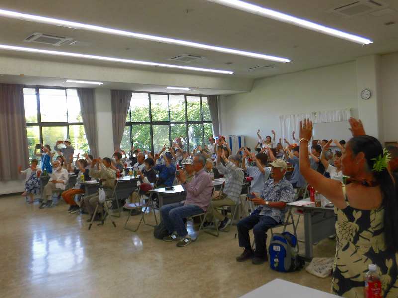 ハワイアンフェスティバルのワークショップに参加する複数の参加者が、講師に合わせて両手を高く上げて楽しんでいる様子を捉えた写真