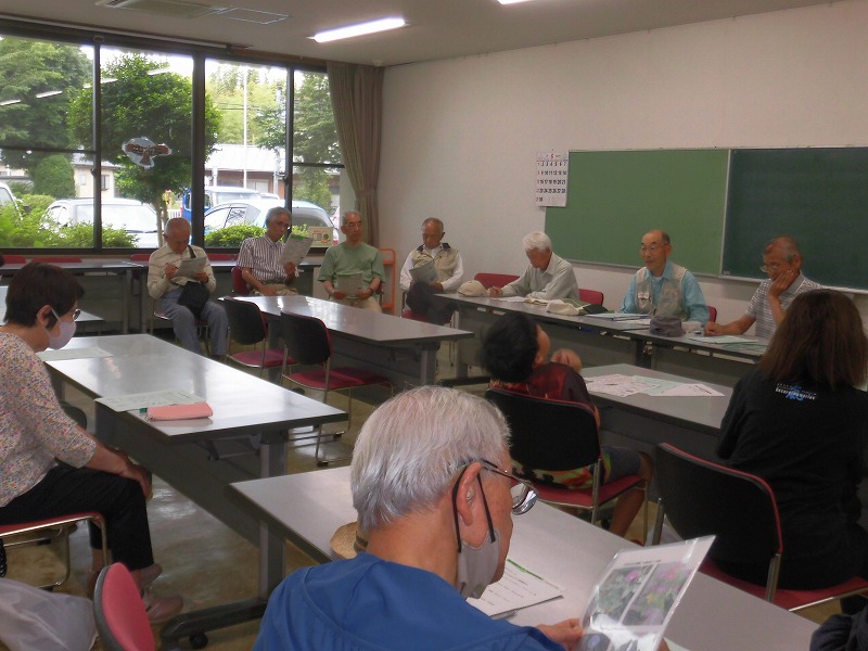 室内で行われている会議で、複数の高齢者がテーブルを囲んで座り、手元の資料に目を通しながら話し合いに参加している様子を捉えた写真