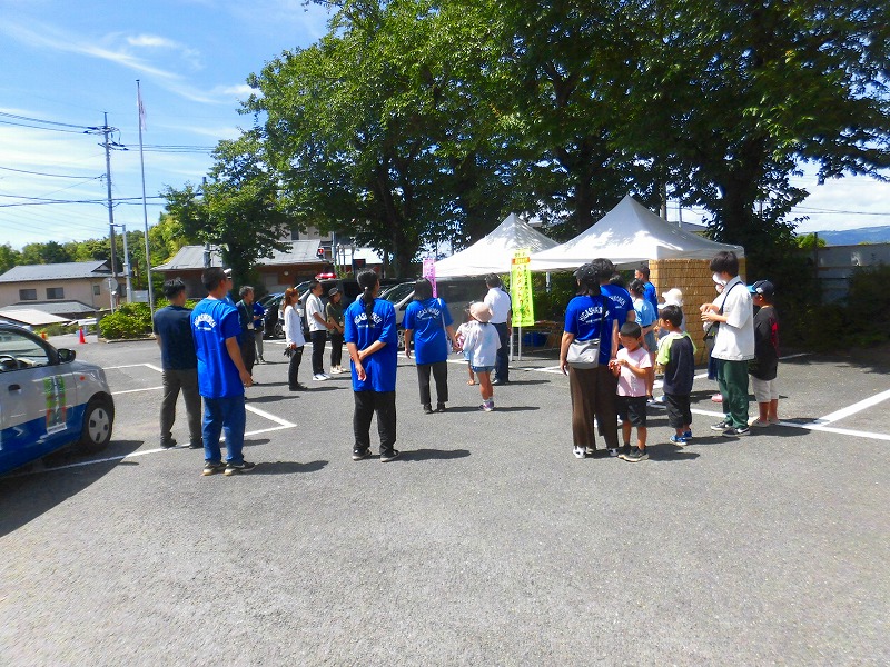 快晴の空のもと、駐車場に設置された2つの白いテントの周りで、青いTシャツを着たスタッフと、子どもを連れた家族が楽しそうに交流している屋外イベントの写真