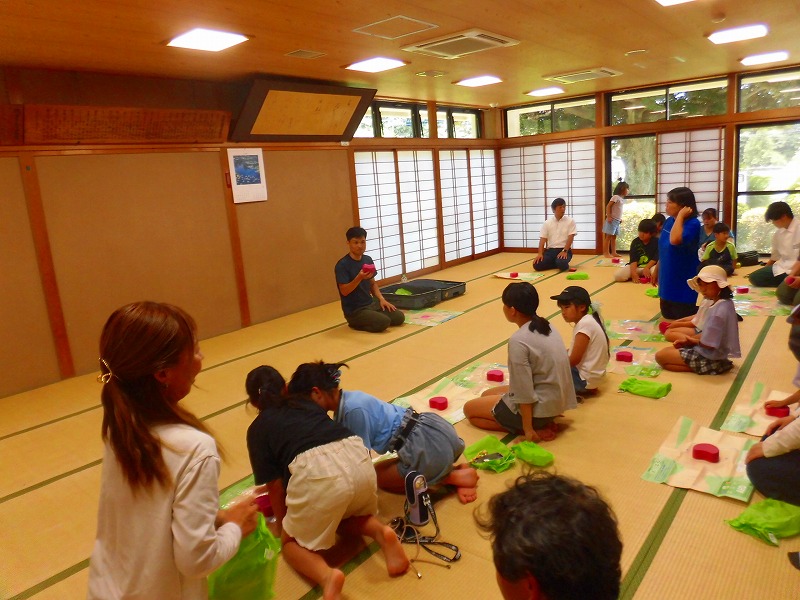 和室の畳の上で、大人と子どもたちが指導者を囲むように座り、男性から心肺蘇生訓練を受けている講習会の様子を捉えた写真