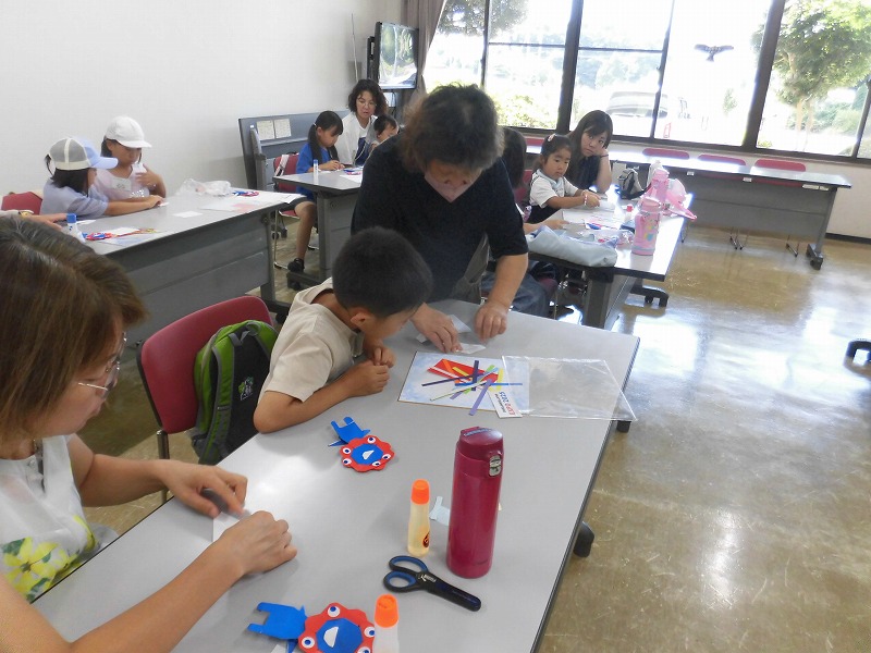 山口みどりさんが、紙とハサミを使って工作をしている男の子に優しくアドバイスを送り、周囲の大人と子どもたちもそれぞれ制作に取り組んでいる写真