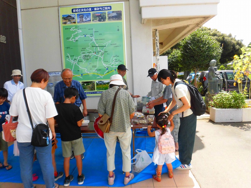 施設の入り口に掲示された地図の前で、大人たちが子どもたちに何かを配っており、多くの人が集まっている屋外イベントの写真