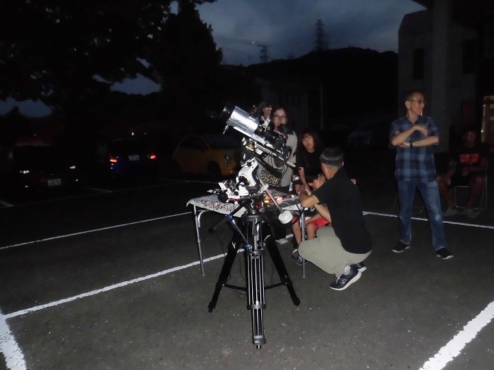 薄暗い屋外で、大人と子どもが天体望遠鏡を覗き込んでいる様子を捉えた写真