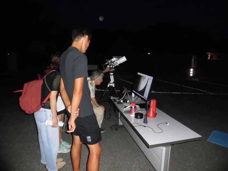 暗い屋外の地面に設置された天体望遠鏡のそばで、モニターに映し出された月を見ながら、複数の人々が夜空の観測を楽しんでいる写真