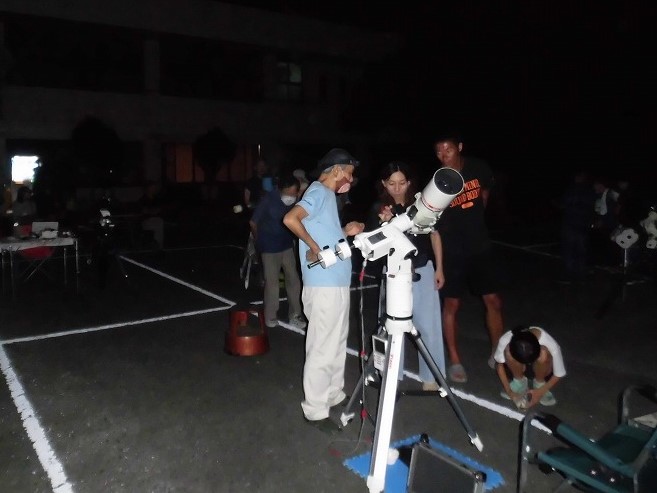 夜の暗闇の屋外で、数人の大人と子供が、三脚に設置された天体望遠鏡を覗きながら楽しんでいる様子を捉えた写真
