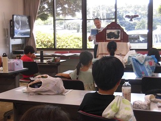 室内で横溝さんが紙芝居を実演しており、テーブルに座った複数の子供たちがその紙芝居を熱心に見つめている様子の写真