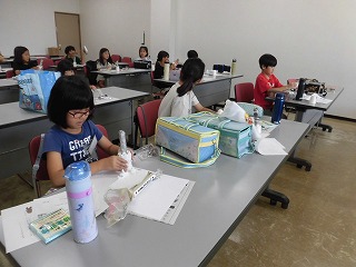 複数の机が並ぶ室内で、小学生の子どもたちが工作道具や教材を広げて、熱心に作業に取り組んでいる様子の写真