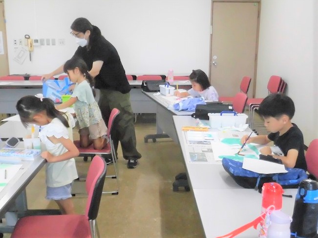 広々とした室内に並べられた机で、子どもたちが各自の道具を使い、高橋智也さんのサポートを受けながら絵画や工作に熱心に取り組んでいる写真