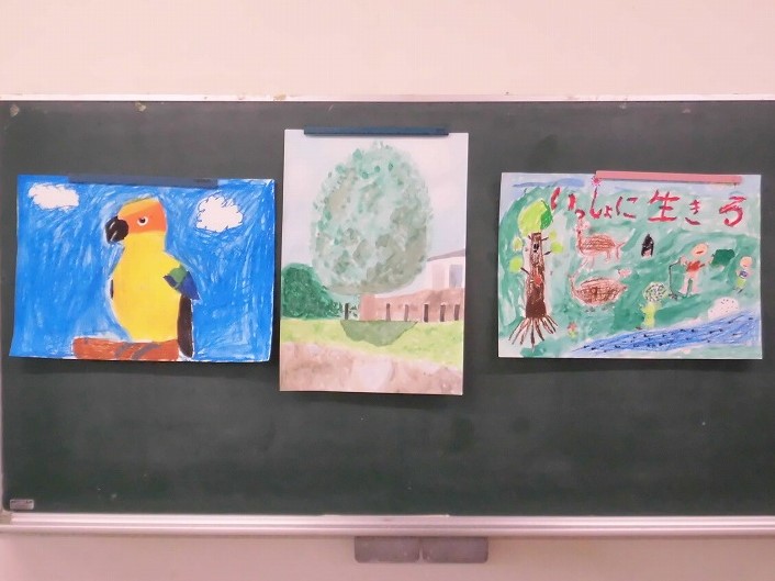 黒板に3枚の絵が掲示されており、左から青い空と黄色いオウム、木のある風景、「いっしょに生きる」と書かれた動物や人間が描かれている作品が並んでいる写真