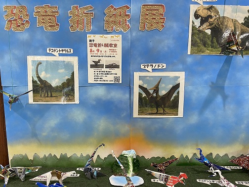 「恐竜折り紙教室」と書かれた案内が掲示され、ティラノサウルスやプテラノドンなどの恐竜の写真や折り紙作品が展示されている写真