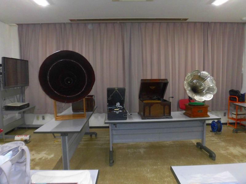 屋内展示室に設置された複数の長机の上に、大きなラッパの蓄音機や木製のボックス型蓄音機など、様々な種類のアンティークな蓄音機が展示されている写真