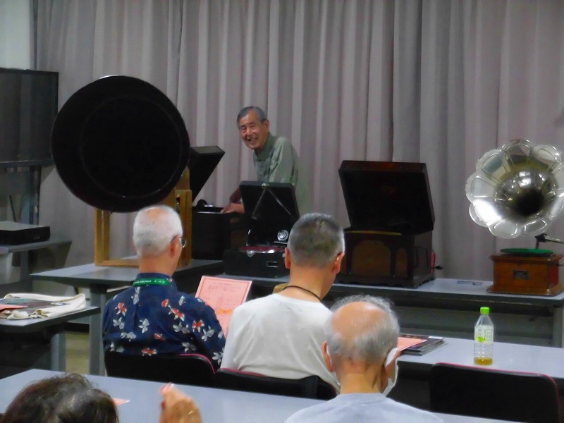 複数の机が並んだ室内で、男性が蓄音機を操作しており、それを鑑賞するために集まった人々が熱心に見守っている様子を捉えた写真