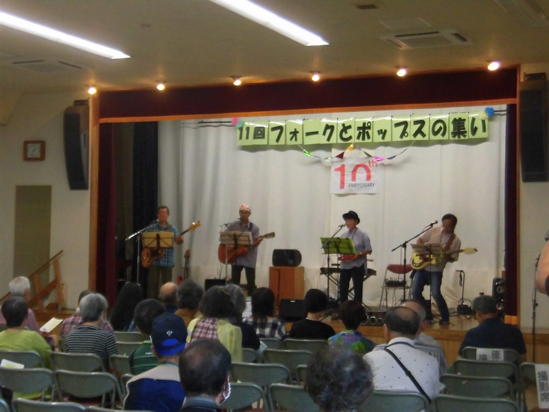屋内のステージに設置された「11回フォークとポップスの集い」と書かれた横断幕の下で、複数の男性がギターやベースを演奏しており、多くの観客が椅子に座って音楽を聴いている写真