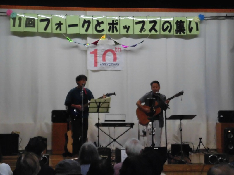 「11回フォークとポップスの集い」という横断幕が掲げられたステージで、2人の男性がそれぞれアコースティックギターとエレキギターを演奏し、観客がそれを見つめている音楽イベントの写真