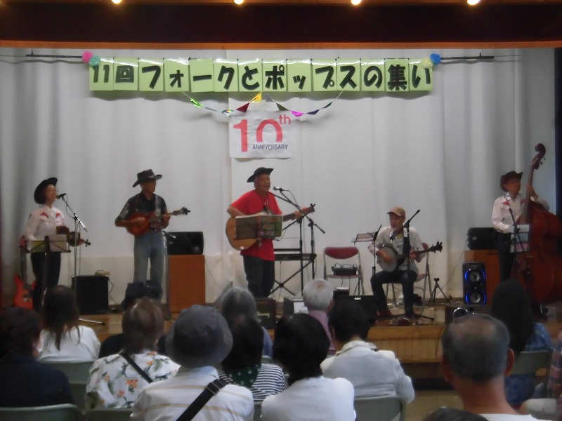 「11回フォークとポップスの集い」という横断幕が掲げられたステージで、赤いシャツやカウボーイハットを身につけたバンドメンバーが、アコースティックギターやウッドベースを演奏し、観客がそれを見つめている写真