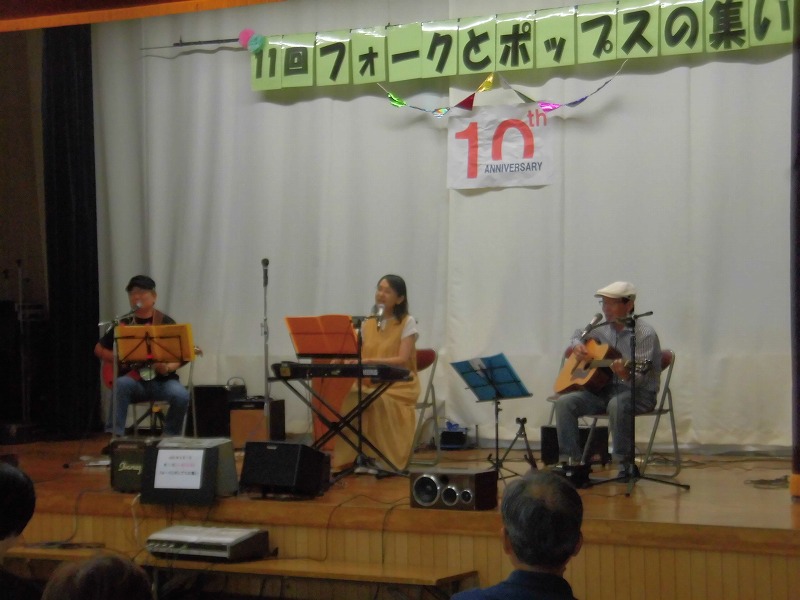 「11回フォークとポップスの集い」という横断幕が掲げられたステージで、女性がキーボードを演奏しながら歌い、両隣に座った男性がギターを弾いている音楽ライブの写真