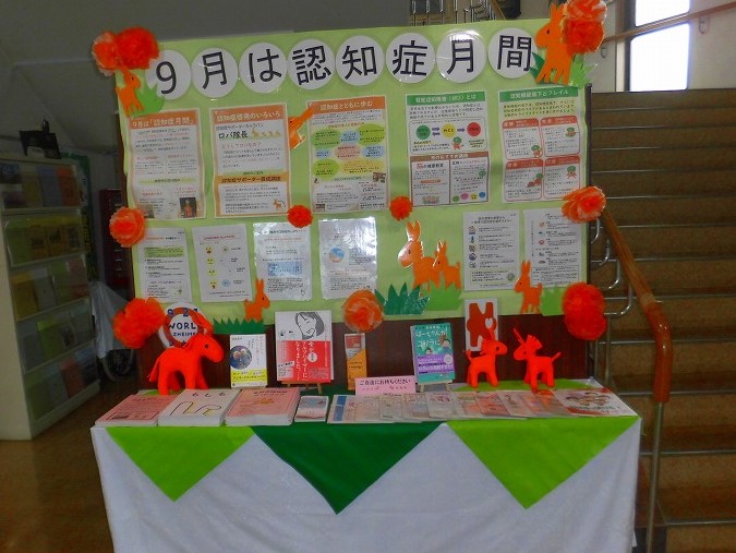 「9月は認知症月間」と書かれた緑色の掲示板が設置され、その前には認知症に関するパンフレットや書籍、そしてオレンジ色の動物のぬいぐるみが飾られている写真