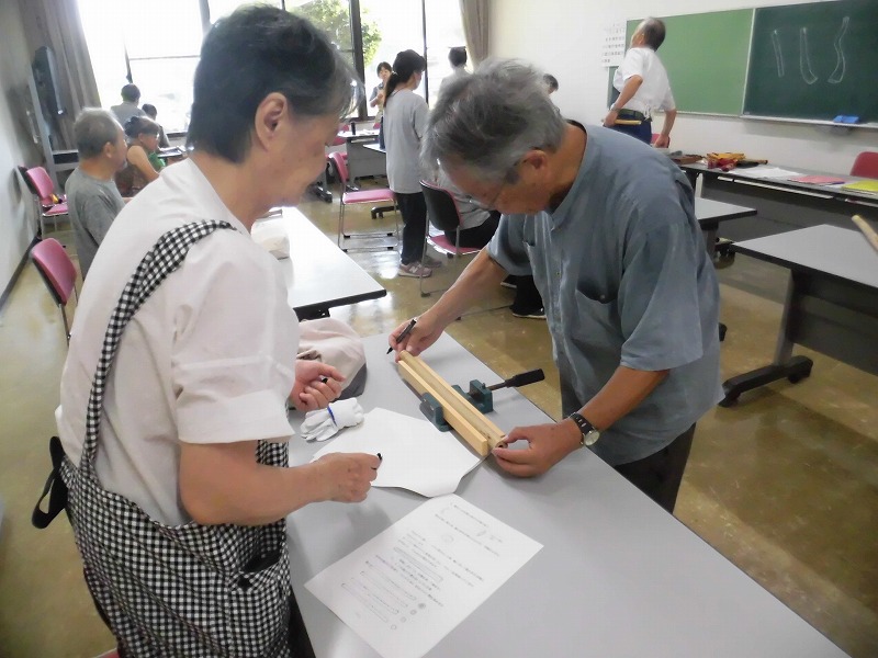 白と水色のシャツを着た男女二人が、教室内の机の上に置かれた木製の細工物に定規を当てて印をつけている様子を、他の参加者や奥の黒板とともに捉えた木工教室の写真
