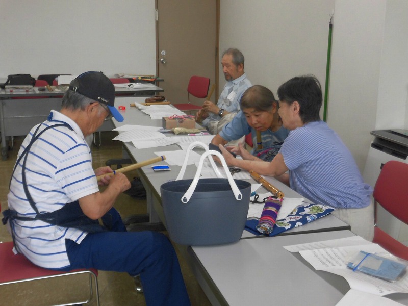 男女4人が長机に向かい、手前の男性は棒状の木工品を削る作業をしており、他の参加者は話をしながら作業に取り組んでいる様子の写真