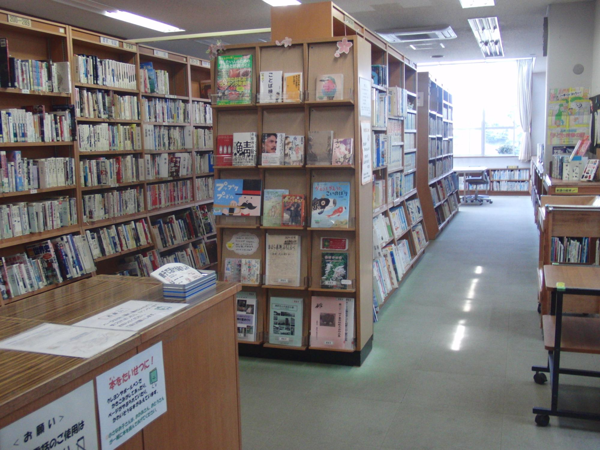 明るい木製の書架が並ぶ図書館の室内で、手前には受付カウンターがあり、その奥には通路を挟んで壁一面の書架と、表紙を見せるように展示された本が並ぶ棚が配置されている写真