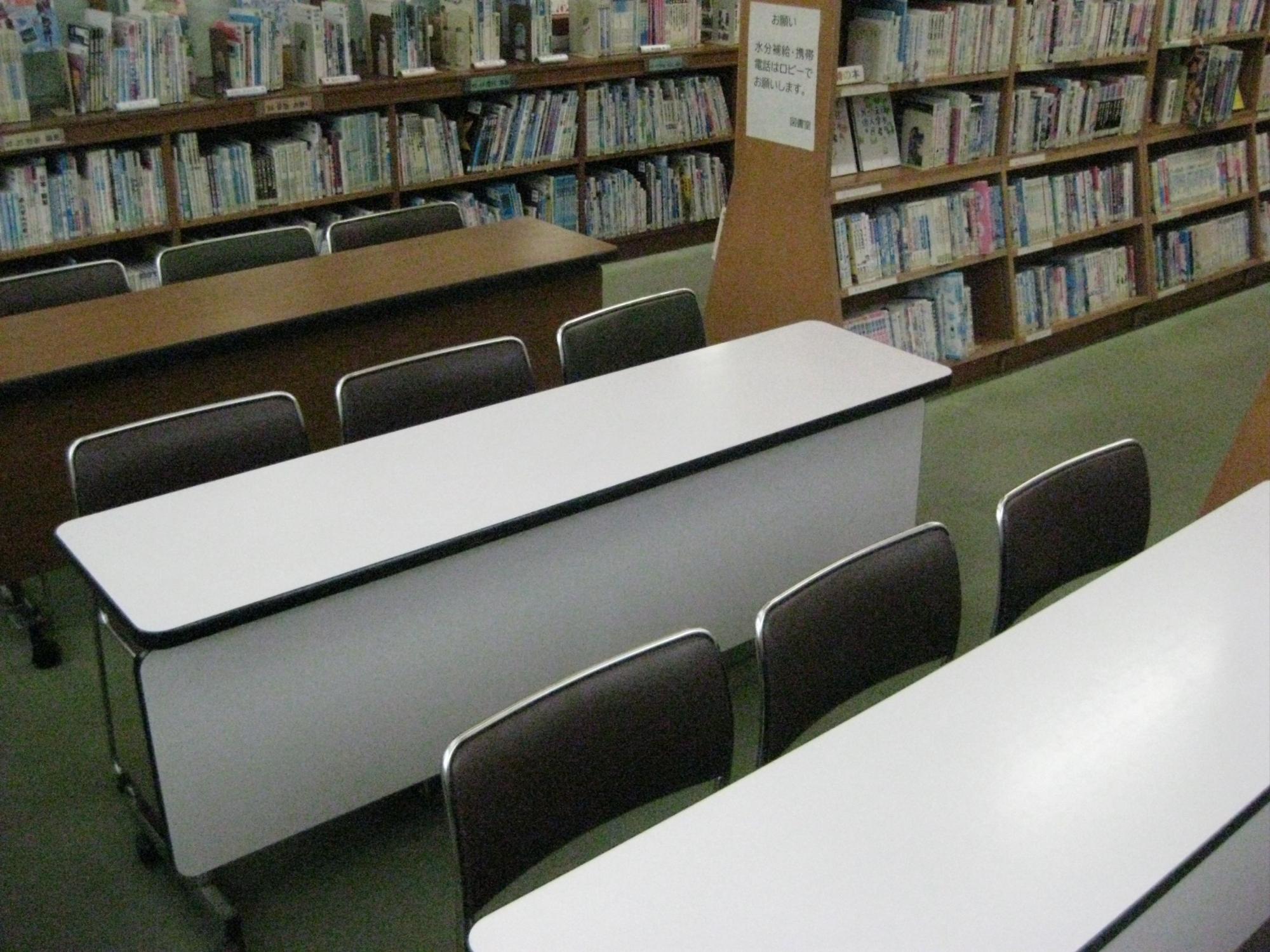 両側に壁一面の書架に本が並び、手前から奥にかけて白い天板と木目調の長机が複数設置され、黒っぽい座面の椅子が整然と並べられている、静かで落ち着いた学習スペースの様子の写真