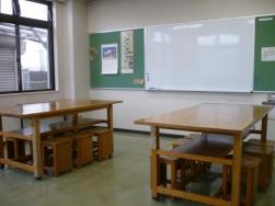 緑色のボードに大きなホワイトボードが設置され、手前には座面が収納できる木製のベンチとテーブルが2組置かれている創作活動室の写真