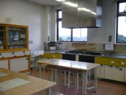 ステンレス製の大きな換気フードが設置され、ガスコンロと流し台がある調理スペースと、手前には作業台となる調理実習用の机が複数置かれている調理実習室の写真