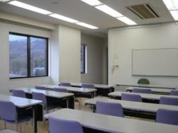 大きな窓があり蛍光灯のある明るい室内で、正面にホワイトボードが設置されており、長机が並んだ会議室の写真