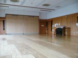 木目調の床で、右奥には黒いアップライトピアノが置かれている、広々とした多目的ホールの室内写真