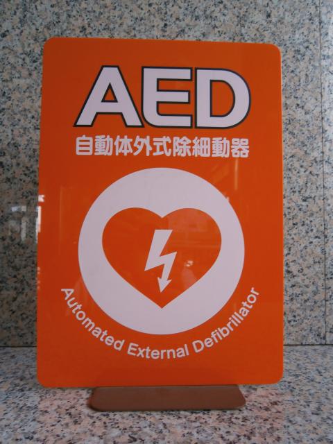 日本語と英語で「AED(自動体外式除細動器)」と書かれており、中央にハートと稲妻のマークが描かれた、オレンジ色のAED設置場所を案内する表示板の写真