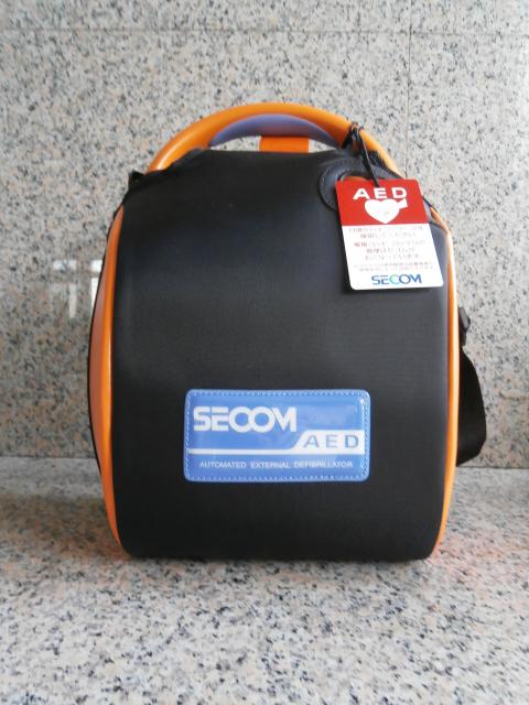 セコムのロゴと「AED」の文字が記された、黒とオレンジ色の専用ケースに収納された自動体外式除細動器を正面から撮影した写真