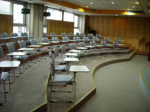 複数の机と椅子が半円状に階段状に並べられており、前方のステージに向かって配置されている、木目調の壁が特徴的な音楽室を捉えた写真