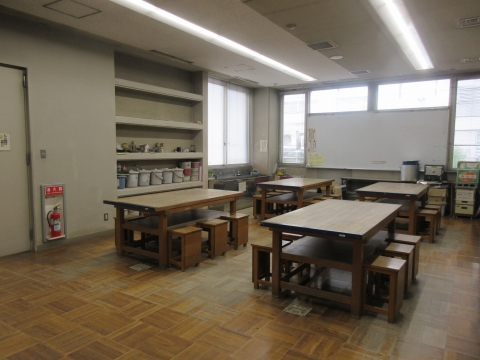 広い空間に、複数の木製の大きな作業台と小さな椅子が並べられており、奥には白い黒板と棚が配置されている創作活動室の写真