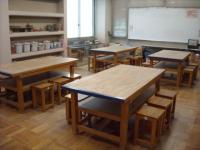 広い空間に、複数の木製の大きな作業台と小さな椅子が並べられており、奥には白い黒板と棚が配置されている創作活動室の写真